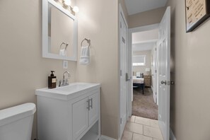 Une salle de bain avec un meuble-lavabo blanc, un miroir avec éclairage intégré, des toilettes et un couloir menant à une chambre.