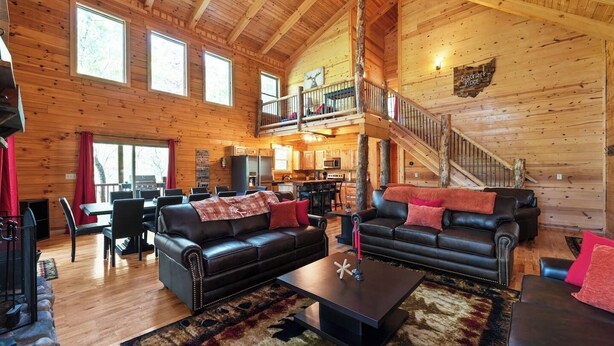 A spacious log cabin interior with a dining area, two black leather sofas, a coffee table, and a staircase leading to an upper level. - Spacious | Sleeps 22 | Pool Table, EV Charger,King (Logan)