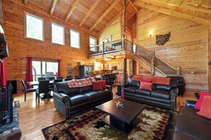 A spacious log cabin interior with a dining area, two black leather sofas, a coffee table, and a staircase leading to an upper level. - Spacious | Sleeps 22 | Pool Table, EV Charger,King (Logan)