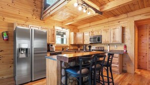 A wooden cabin-style kitchen with a central island, stainless steel appliances, and bar stools. - Spacious | Sleeps 22 | Pool Table, EV Charger,King (Logan)