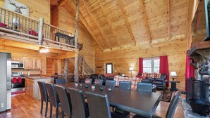 A wooden cabin interior with a dining area, kitchen, and a fireplace. - Spacious | Sleeps 22 | Pool Table, EV Charger,King (Logan)