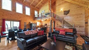 A spacious lodge with a high ceiling, wooden walls, a dining area, a living room with leather sofas, and a staircase leading to an upper level.