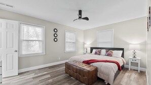 A bedroom with a bed, a ceiling fan, a window with blinds, a bedside table with a lamp, and a wooden floor. - Pool Table • Fire Pit • Bar • King • Sleeps 22 (Galena)