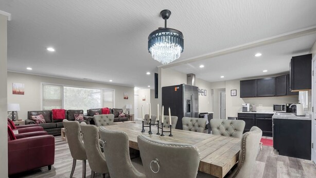 A spacious living area with a dining table, chairs, a chandelier, a kitchen with dark cabinets, and a large window. - Pool Table • Fire Pit • Bar • King • Sleeps 22 (Galena)