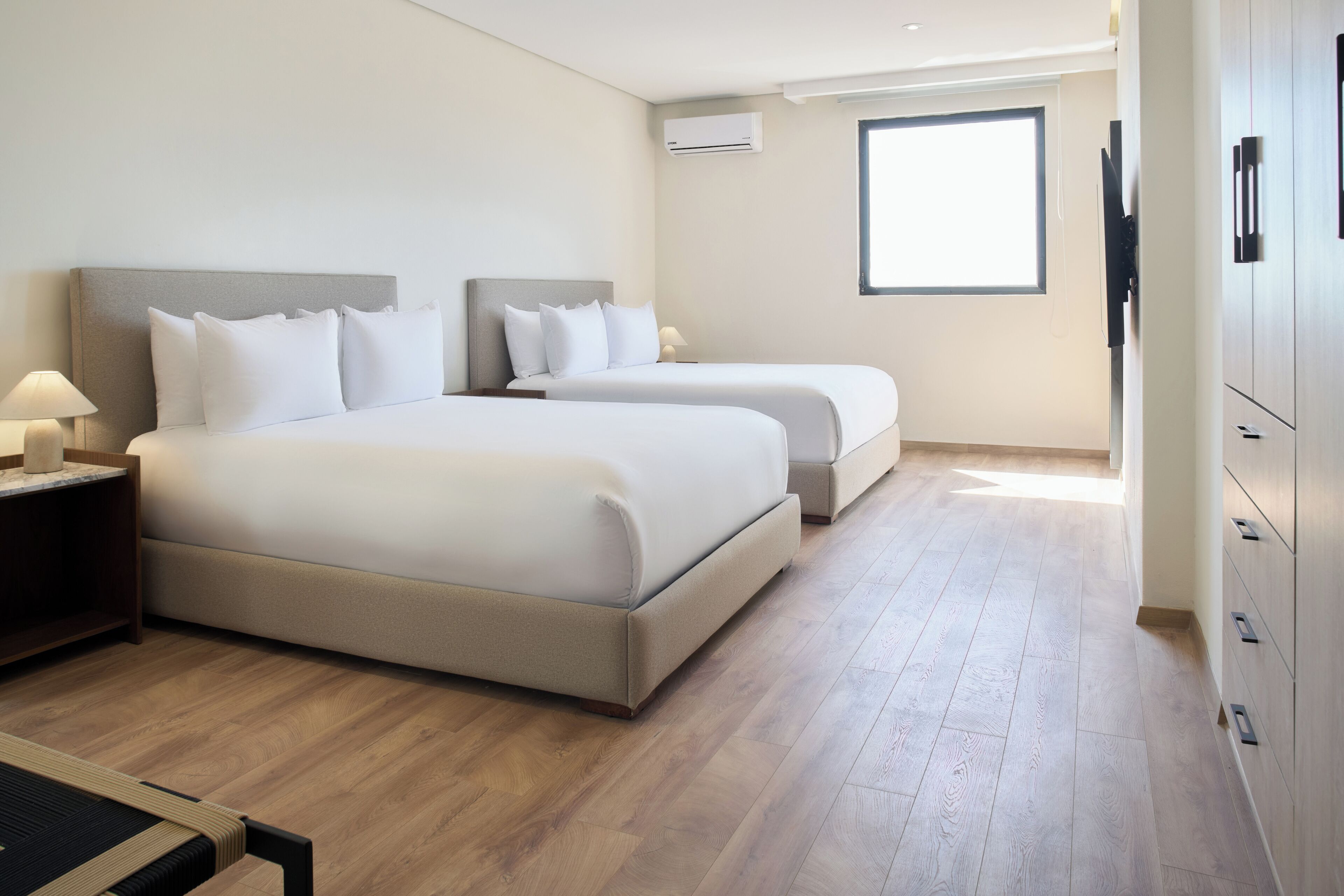 A hotel room with two beds, a wooden floor, a window, and a TV on the wall.