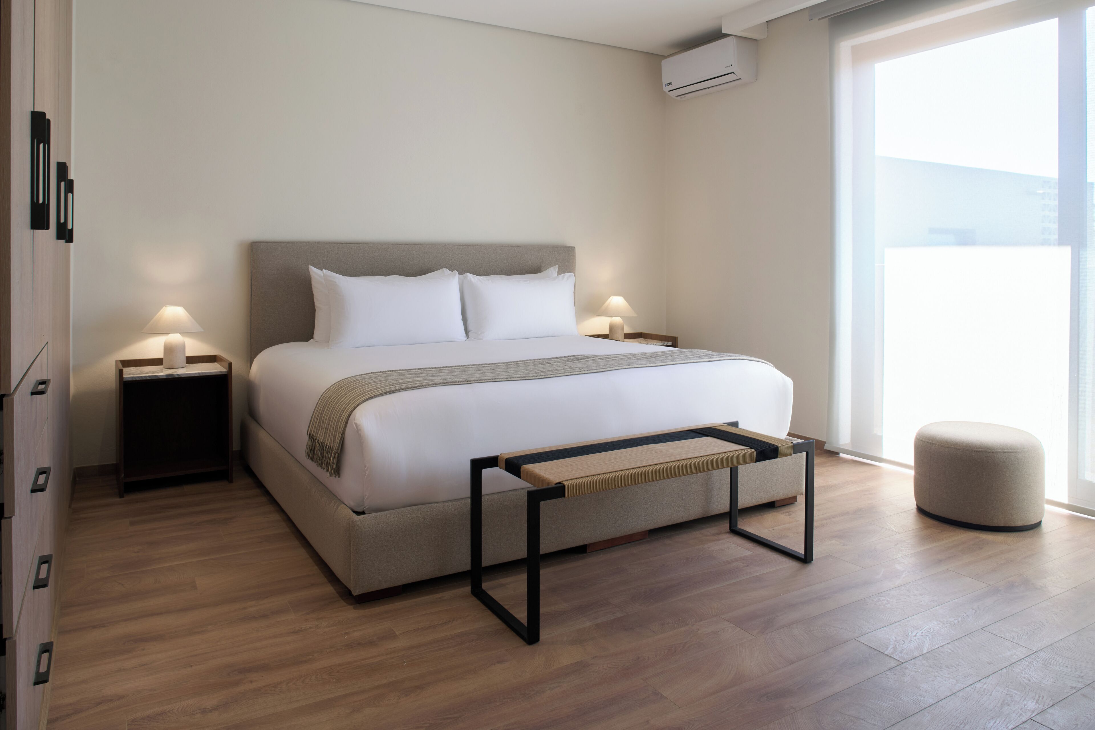 A modern bedroom with a large bed, two bedside tables with lamps, a bench, and a view of the outdoors.