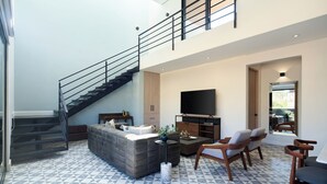 Uma sala de estar moderna com um sofá seccional cinza, uma mesa de centro e duas cadeiras de madeira. Uma televisão de tela plana está instalada na parede, e uma escada com corrimão preto leva a um andar superior.