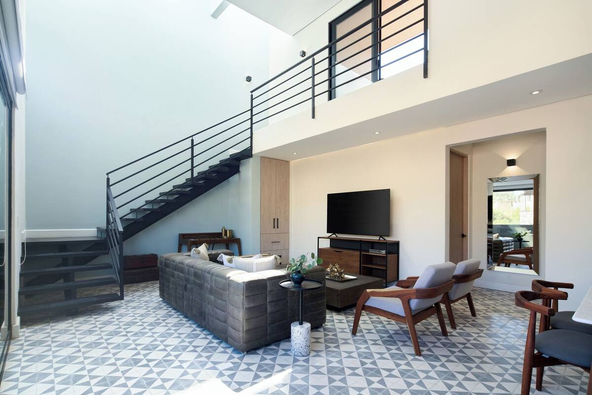 Ein modernes Wohnzimmer mit einem grauen Ecksofa, einem Couchtisch und zwei Holzstühlen. Ein Flachbildfernseher ist an der Wand montiert, und eine Treppe mit schwarzer Geländer führt auf eine erhöhte Ebene.