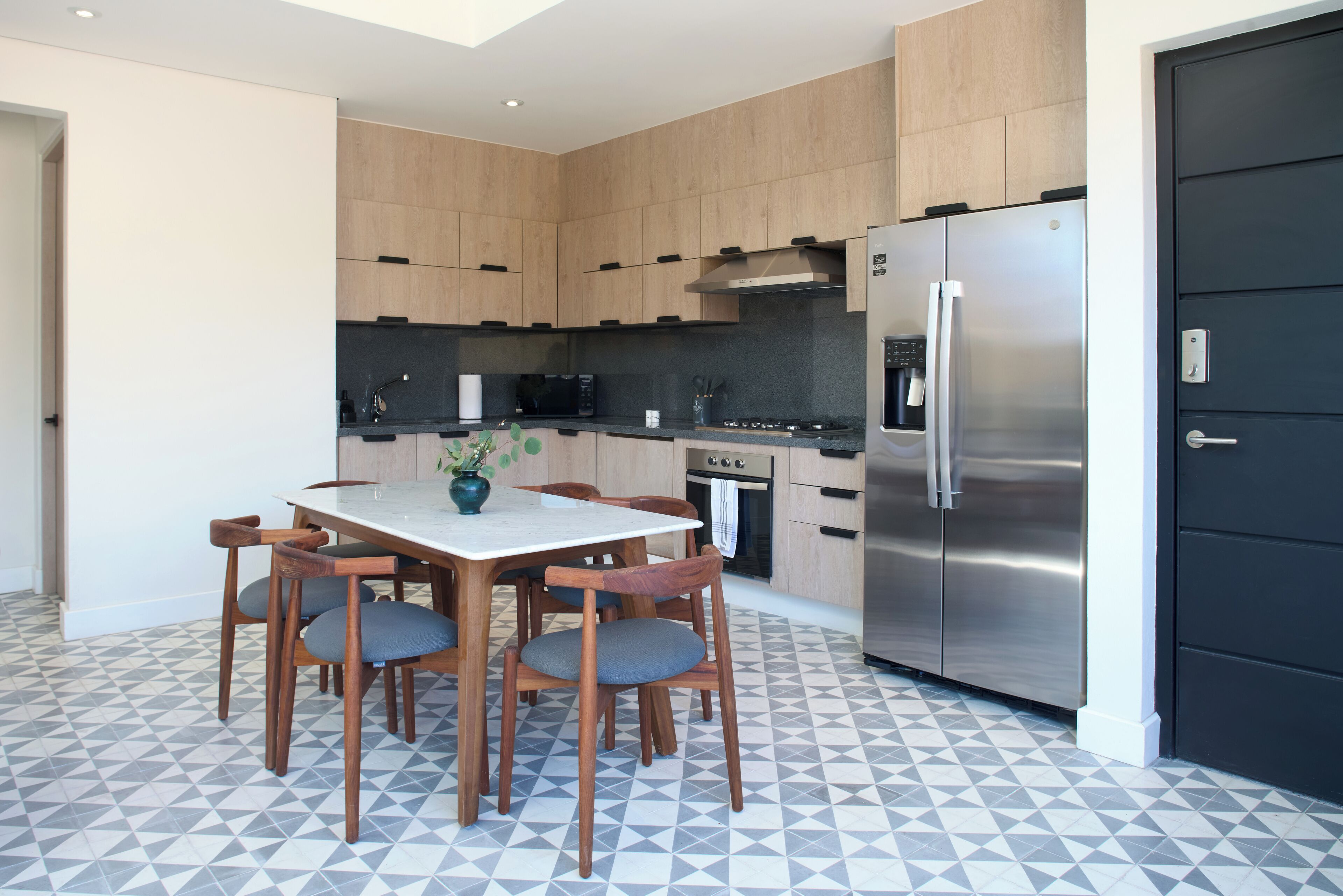 A modern kitchen with wooden cabinets, a stainless steel refrigerator, a dining table with chairs, and a patterned floor.