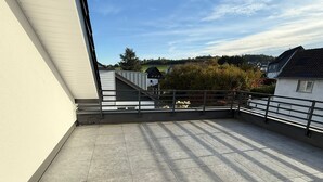 A spacious balcony with tiled flooring, a metal railing, and a view of a residential area with houses and trees. - StayEasy Accommodation (Gummersbach)