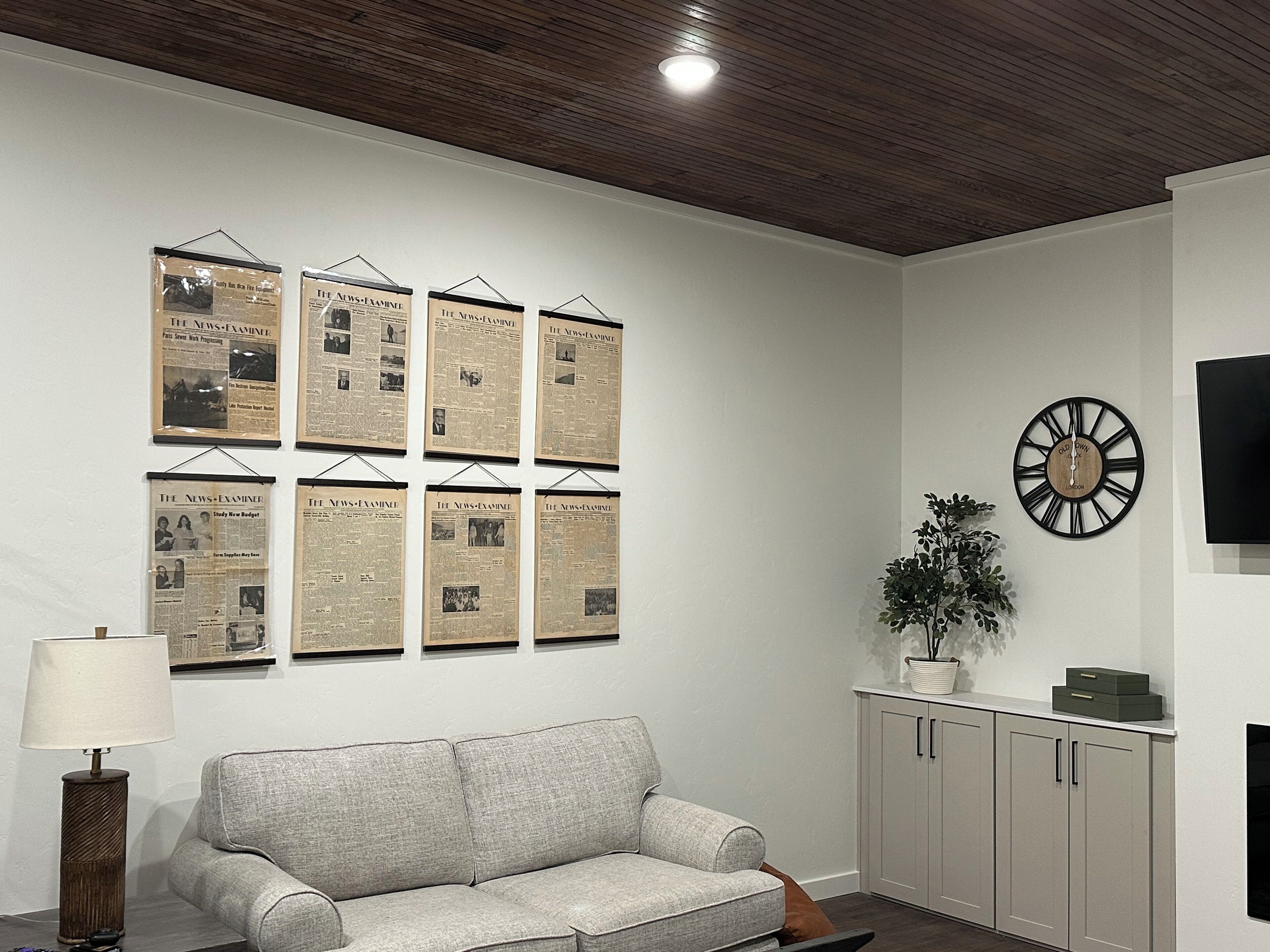 Una sala de estar con un sofá gris, un reloj en la pared y recortes de periódicos enmarcados.
