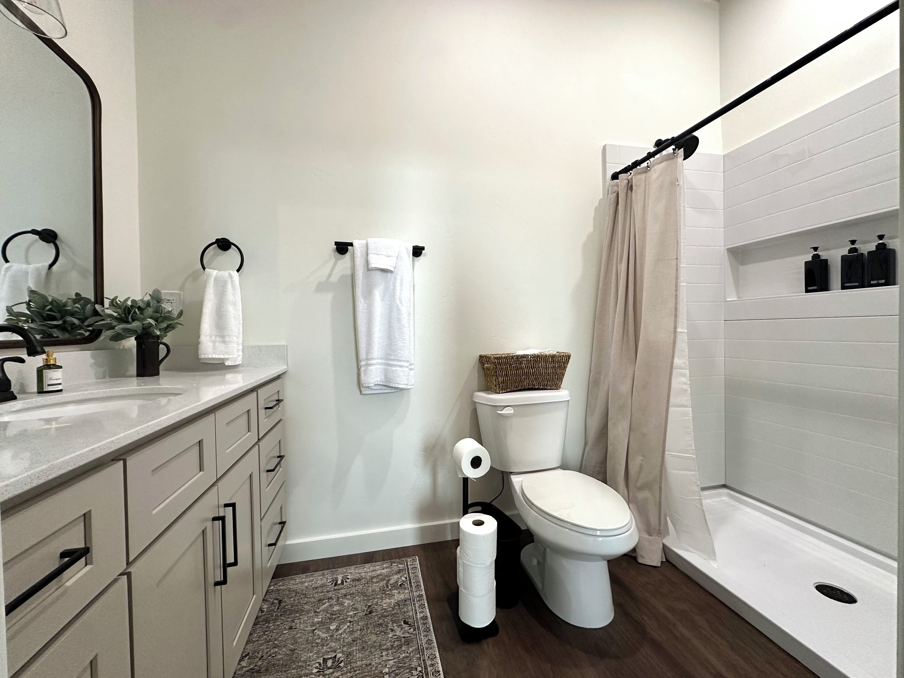 A bathroom with a toilet, sink, and shower. There are towels hanging on hooks, a basket on the toilet, and toiletries on the wall.