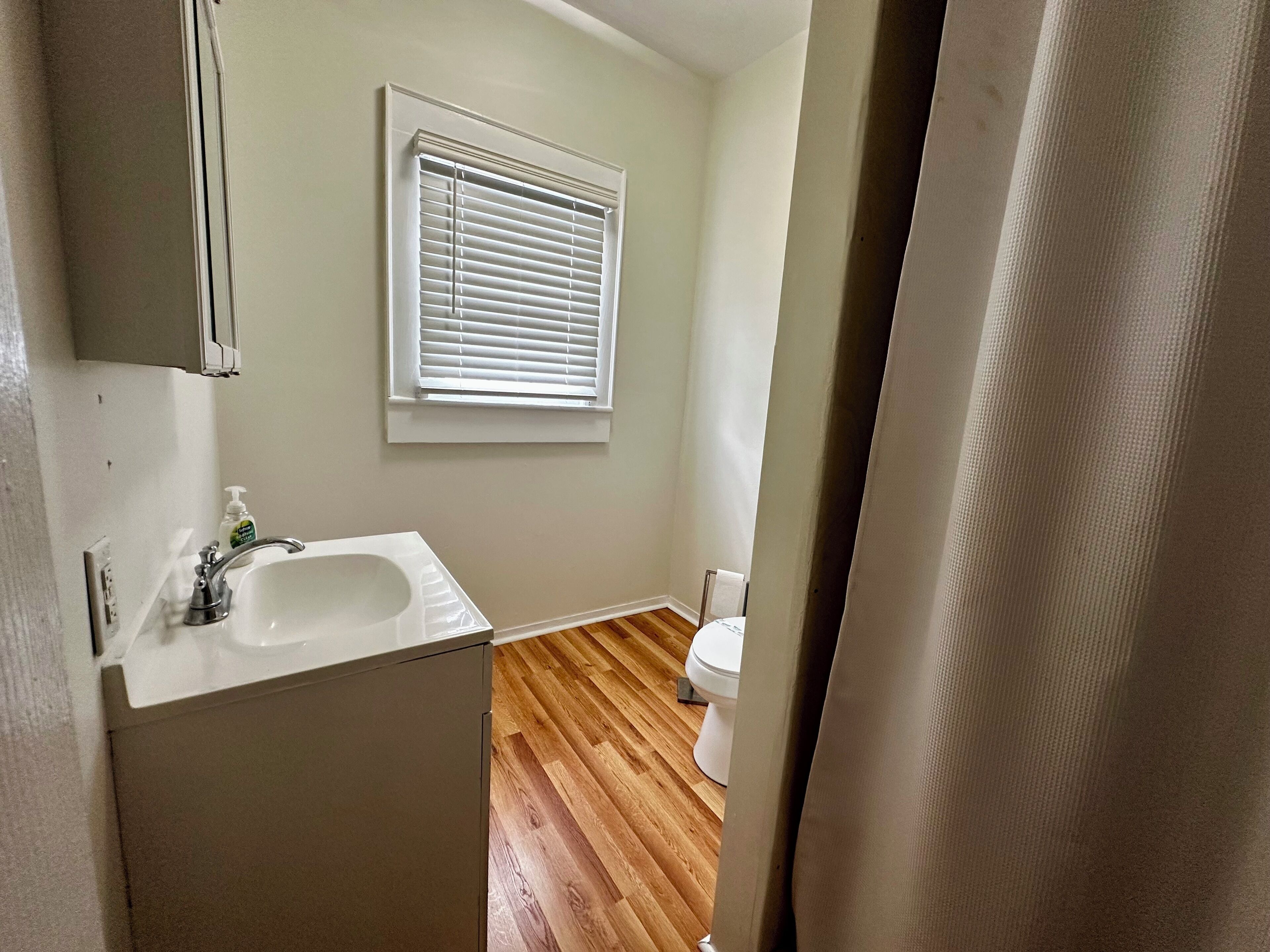 A bathroom with a white sink, a window with blinds, a toilet, and wooden flooring.