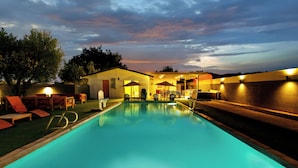 A swimming pool with lounge chairs and umbrellas, surrounded by a building with a brick wall. - The Clubhouse: Sleep 20+, Pool, Pickleball, Hot Tub, Outdoor Movie, Arcades (Indio)