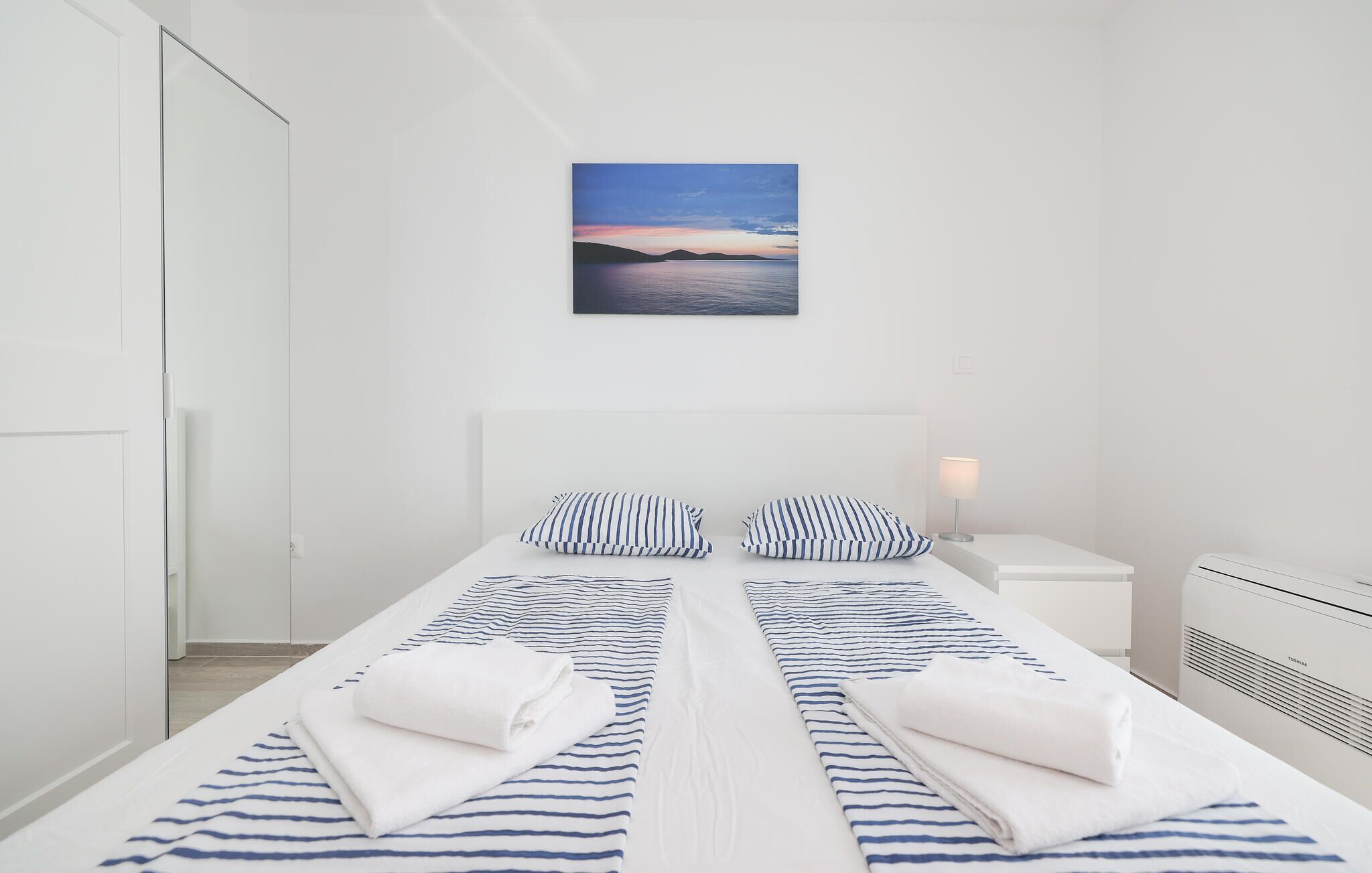 Habitación de hotel con cama, ropa de cama a rayas, dos almohadas, lámpara de noche y un cuadro en la pared de una puesta de sol sobre el agua.