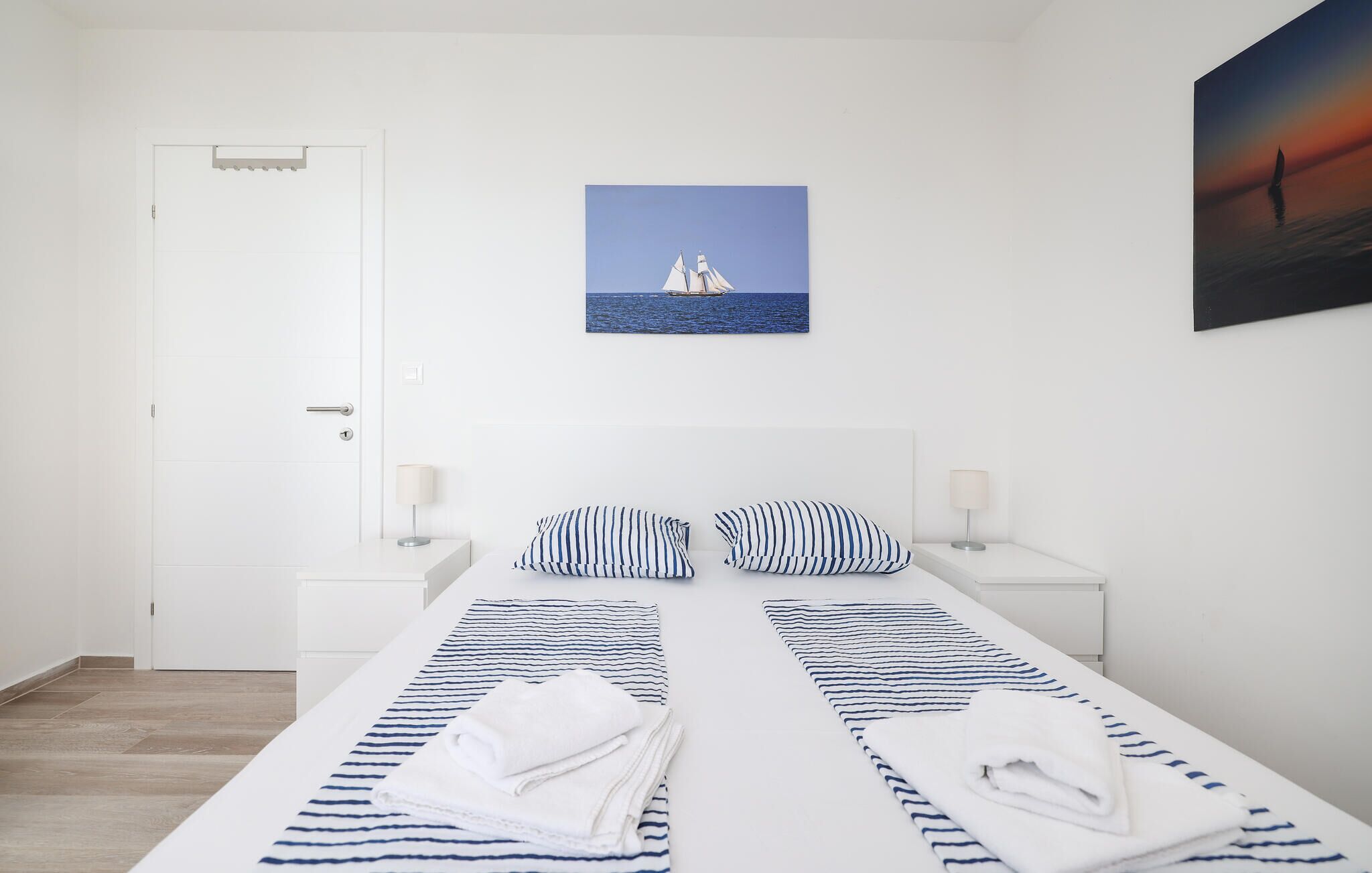 A hotel room with a bed, two bedside tables, a door, and a painting of a sailboat on the wall.