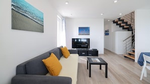A modern living room with a grey sofa, a flat-screen TV, and a coffee table. There is a staircase with a wooden finish and a white railing.
