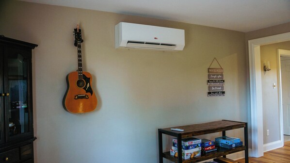 A room with a guitar, an air conditioner, a wall-mounted sign with house rules, and a table with board games.