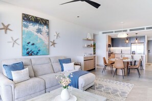Uma sala de estar moderna com sofá, mesa de centro e uma pintura de uma cena de praia. Há uma área de jantar com cadeiras e uma mesa.