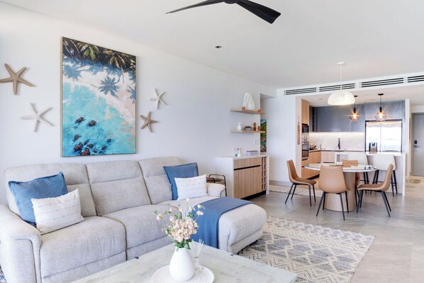 A modern living room with a sofa, coffee table, and a painting of a beach scene. There is a dining area with chairs and a table.