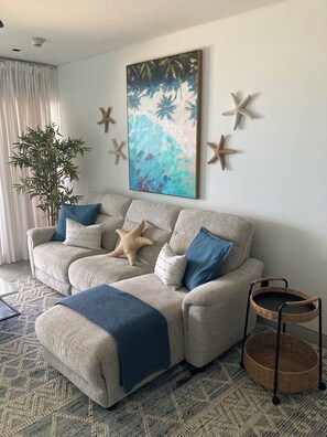 A living room with a beige sofa, blue and white cushions, a starfish decoration, a painting of a beach scene, and a potted plant. - Allure 302: 2BR Beachfront Condo with Infinity Pool, Sun Deck, Gym, Fire Pit etc (Bridgetown)