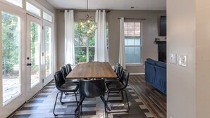 Un coin repas avec une table en bois et des chaises noires, de grandes fenêtres avec des rideaux, et un canapé bleu en arrière-plan.