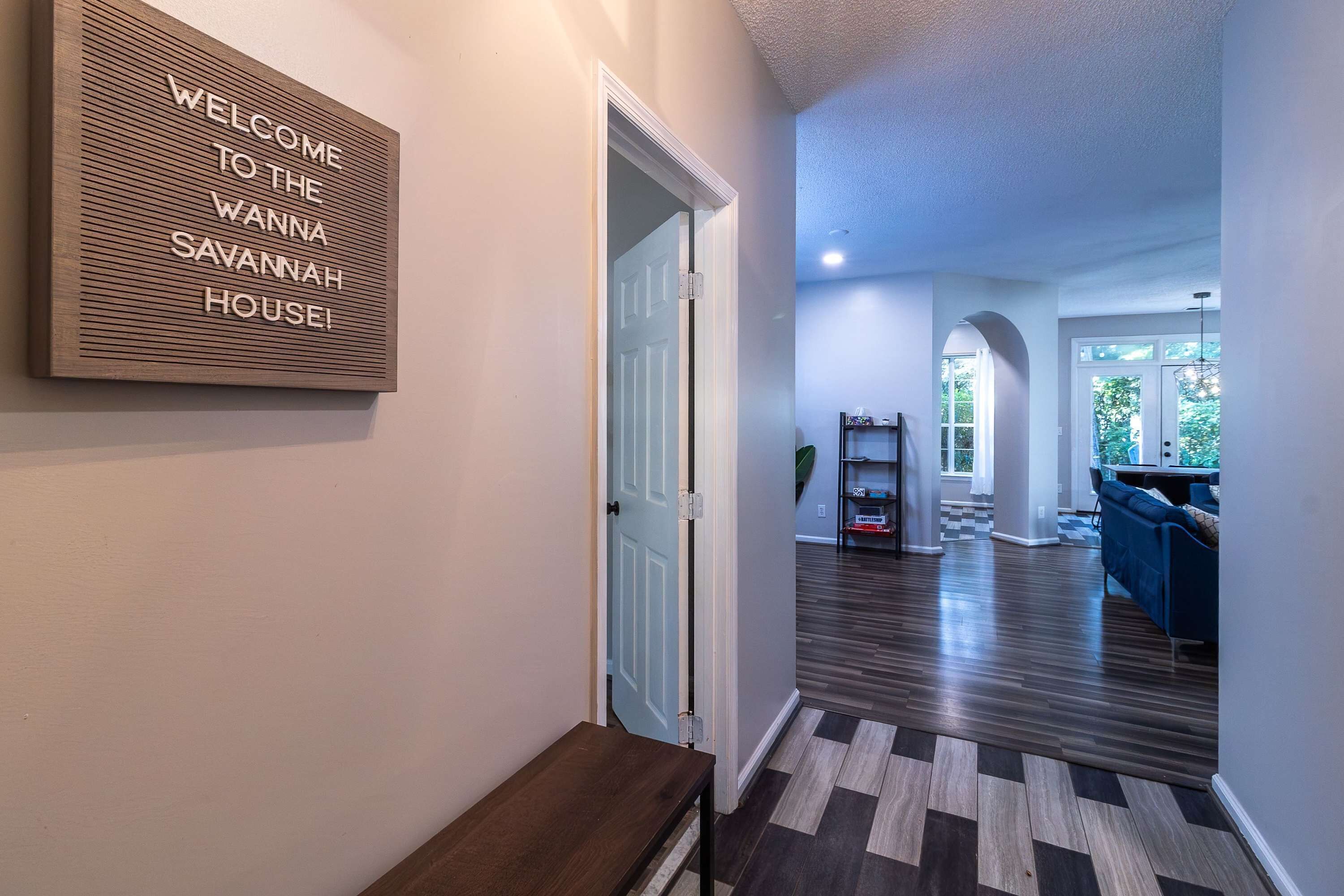 A hallway with a welcome sign, a bench, and a view into a living area with a blue couch and a herringbone-patterned floor.