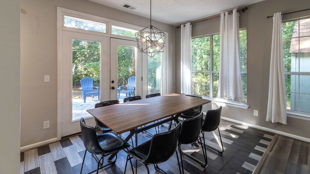 Un espace repas avec une table en bois et des chaises noires, de grandes fenêtres avec des rideaux et un luminaire suspendu au design géométrique.