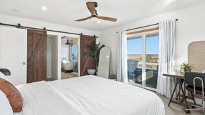Un dormitorio con una cama grande, un escritorio con silla, un ventilador de techo y vistas al desierto a través de una puerta corrediza de vidrio.