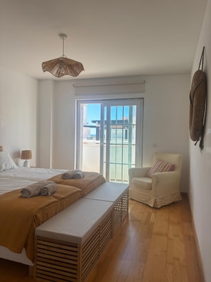 A bedroom with a bed, a bench, a chair, a hanging light fixture, and a view through a glass door.