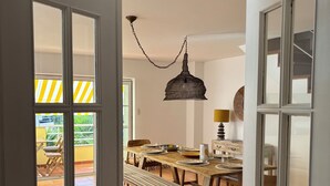 Une porte ouverte donne sur un coin repas où se trouve une table en bois dressée pour quatre personnes, un luminaire suspendu et un banc.