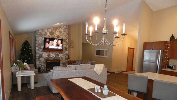 Un salon moderne avec une cheminée en pierre, une grande table à manger en bois et un coin cuisine équipé d’un réfrigérateur et d’un four à micro-ondes.