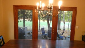Une salle à manger avec une table et des chaises en bois, un lustre, et une vue sur l’espace extérieur aménagé et les arbres, visible à travers des portes vitrées.
