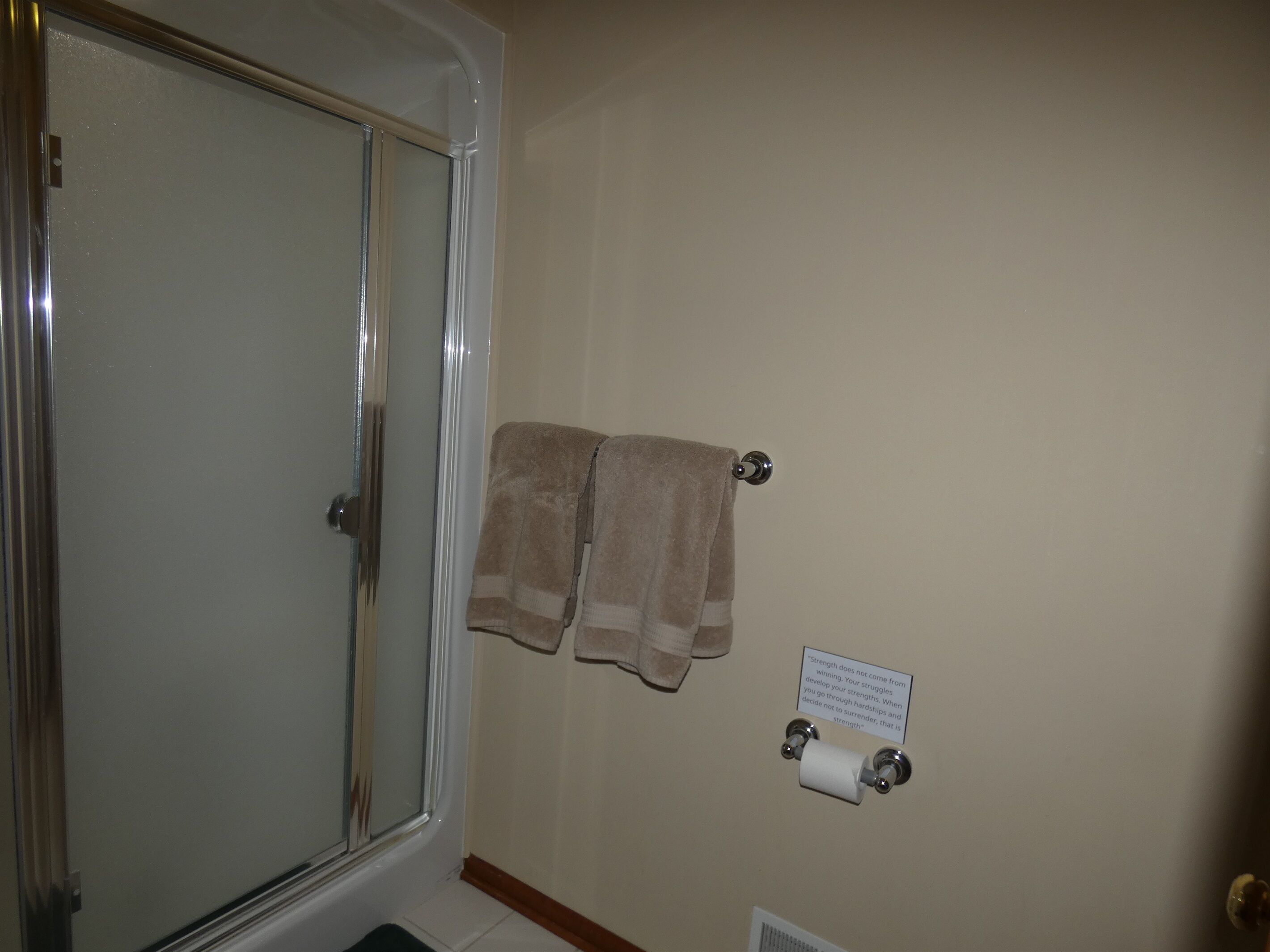 A bathroom with a glass shower door, two towels hanging on a towel rack, and a toilet paper holder with a motivational quote on the wall.