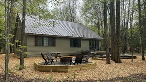 Une cabane avec un toit en métal, entourée d’arbres, et dotée d’un espace extérieur aménagé avec des sièges et d’un foyer.
