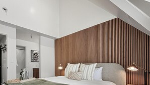 A modern bedroom with a sloped ceiling, a large bed with a green throw, wooden paneling on the wall, and a nightstand with a lamp. - Slope-Side| Modern Remodel | Hot Tub | Park Free (Park City)