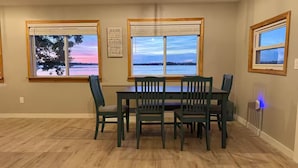 A dining area with a table and four chairs, two windows with views of a body of water, and a wall-mounted sign with text. - Brooko's Lakefront Retreat - Sunset views - 10 Minutes to St. Albans VT (Georgia)