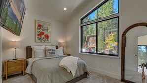 Une chambre avec une grande fenêtre, un lit recouvert d’une couette blanche, une table de chevet, une lampe et une œuvre d’art encadrée accrochée au mur.