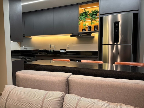 A modern kitchen with dark cabinets, a stainless steel refrigerator, a built-in oven, and a bar area with a seating arrangement.
