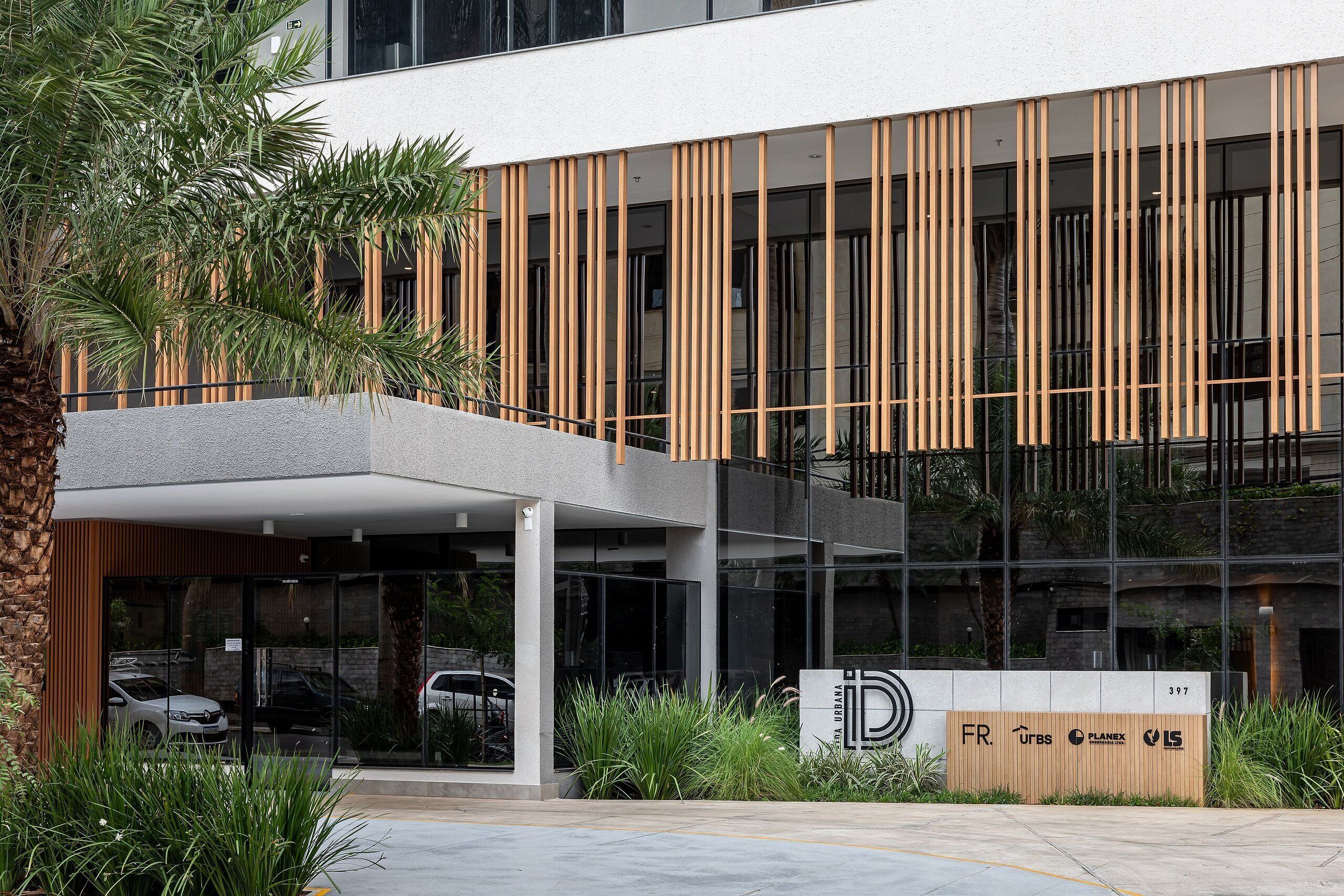 Un edificio moderno con listones verticales de madera, entrada de vidrio y un cartel con varios logotipos.