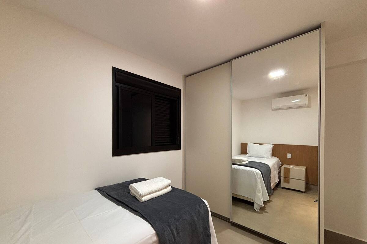 A bedroom with a bed, a bedside table, a mirror, a window with blinds, and an air conditioning unit.