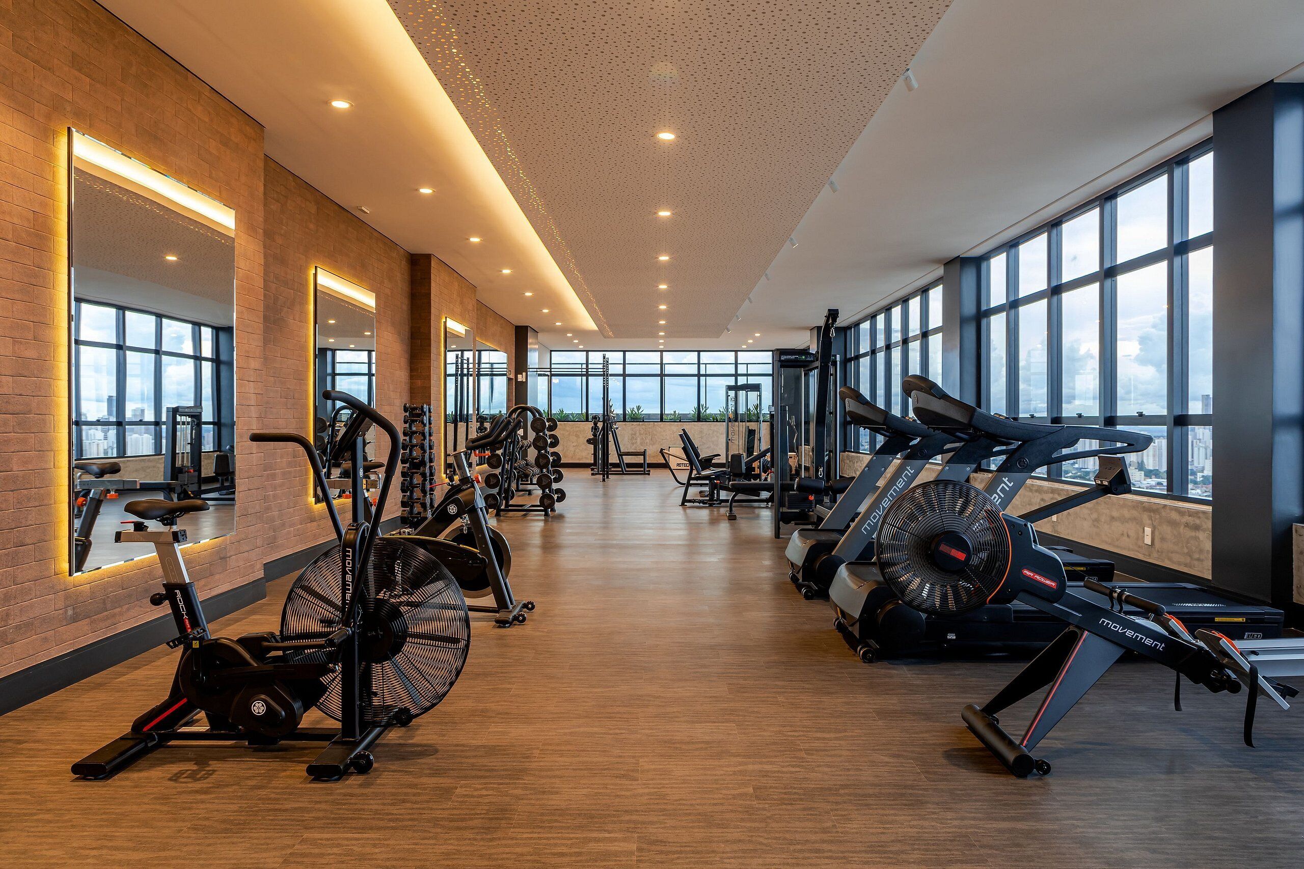 A spacious gym with cardio machines, weightlifting equipment, and large windows offering city views.