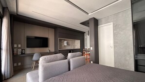 A modern hotel room with a large bed, a flat-screen TV, a seating area with a table, and a large window. - ID Vida Urbana (Goiânia)