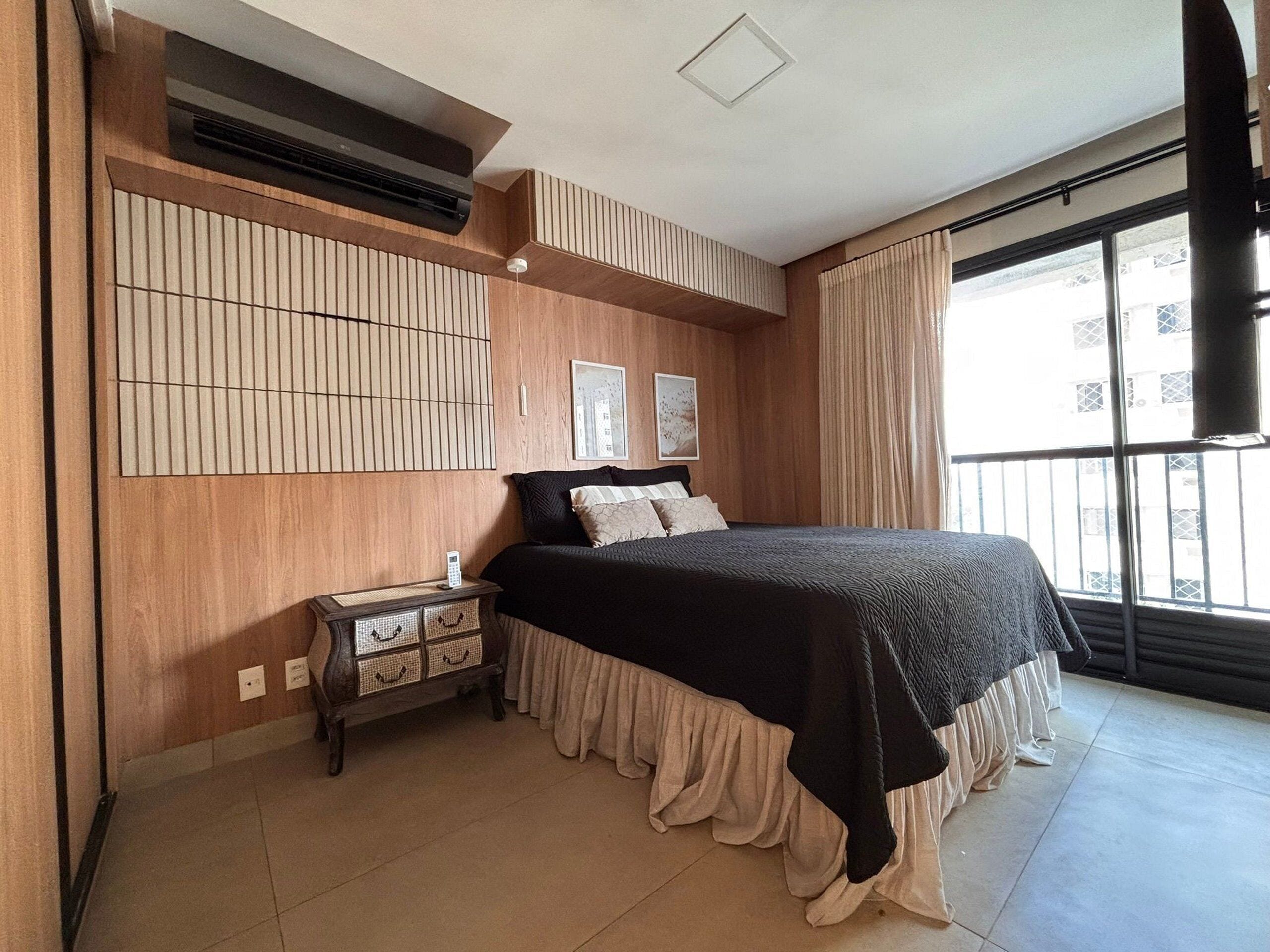 A bedroom with a bed, bedside table, wall-mounted air conditioning unit, and a window with curtains.