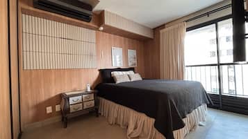 A bedroom with a bed, bedside table, wall-mounted air conditioning unit, and a window with curtains.