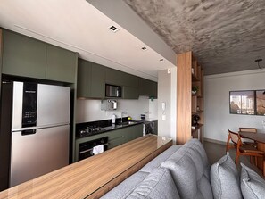 A modern kitchen with a built-in refrigerator, microwave, and stove. There is a wooden bench and a grey sofa in the living area. - ID Vida Urbana (Goiânia)
