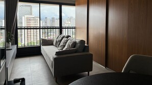 A modern apartment with a large window, a sofa, a dining table, and a view of a cityscape. - ID Vida Urbana (Goiânia)