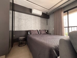 A modern hotel room with a large bed, a small stool, a chair, and a window with curtains. - ID Vida Urbana (Goiânia)