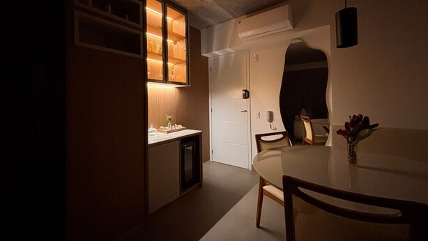 A compact dining area with a round table, chairs, a small kitchen with illuminated shelves, and a white door. - ID Vida Urbana (Goiânia)