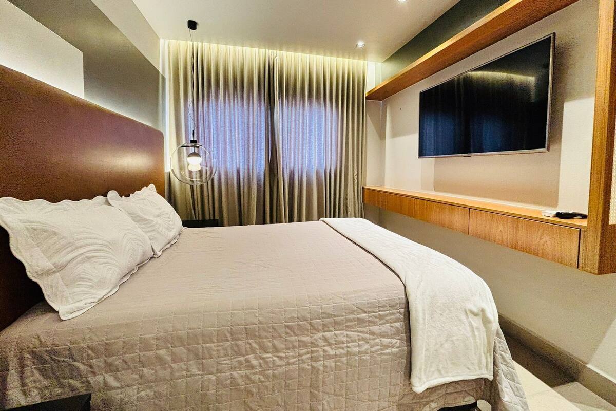 A hotel room with a bed, curtains, a mounted TV, and a bedside lamp.